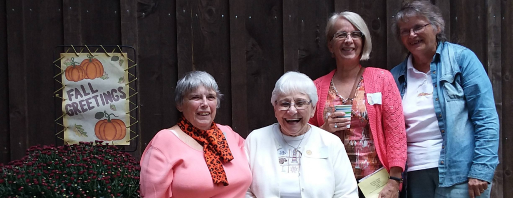 Four smiling women posing outdoors by a wooden fence, fall sign and potted mums on the left side.