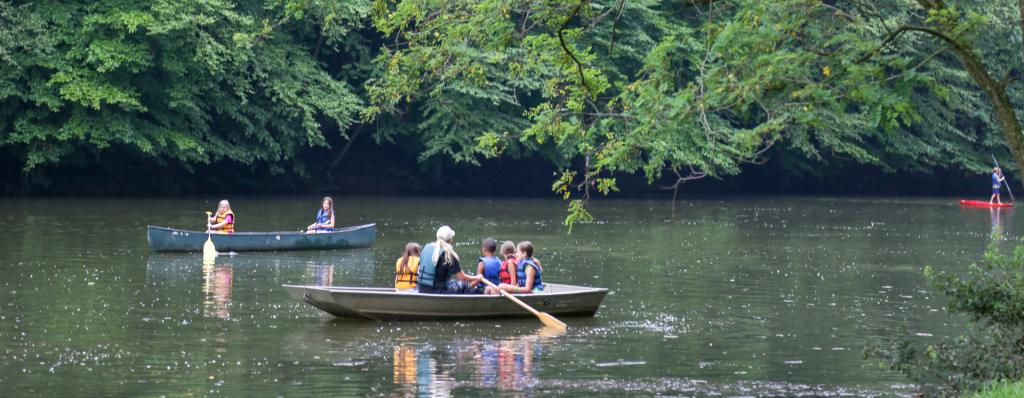 Children in life jackets paddling a canoe on a calm lake, with dense green trees along the shore.