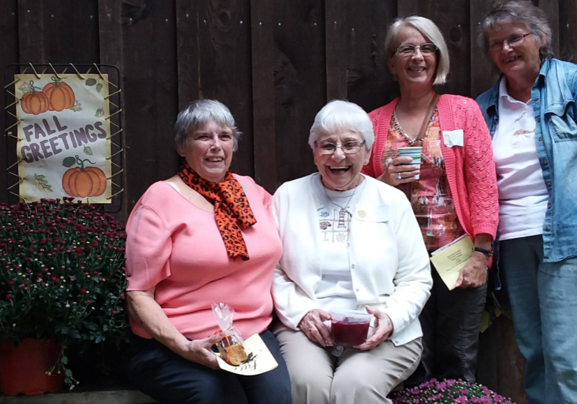 Four smiling women posing outdoors by a wooden fence, fall sign and potted mums on the left side.