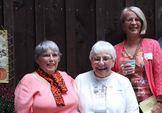 Four smiling women posing outdoors by a wooden fence, fall sign and potted mums on the left side.